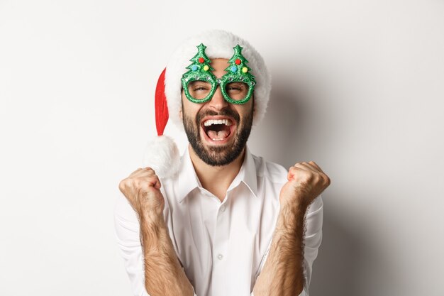 Primer plano del hombre celebrando la Navidad o el año nuevo, con gafas de fiesta de Navidad y gorro de Papá Noel, regocijándose y gritando de alegría, de pie