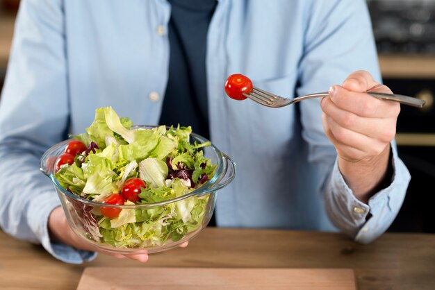 Primer plano de un hombre comiendo el tomate cherry de la ensalada en el recipiente de vidrio con un tenedor