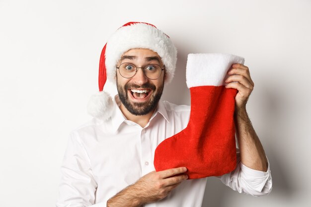 Primer plano de hombre feliz celebrando la Navidad, recibir regalos en calcetín de Navidad y mirando emocionado, con gorro de Papá Noel y gafas