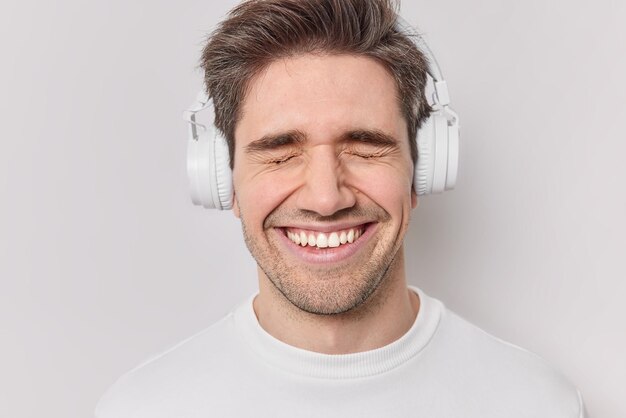 Primer plano de hombre feliz sonríe ampliamente mantiene los ojos cerrados usa auriculares estéreo en los oídos escucha música favorita con sonido fuerte aislado sobre fondo blanco Emociones felices y entretenimiento