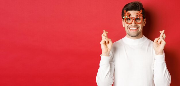 Primer plano de un hombre guapo emocionado, con gafas de fiesta y suéter blanco, cruzando los dedos para tener buena suerte, celebrando la Navidad y esperando el deseo, anticipando regalos, de pie sobre un fondo rojo.