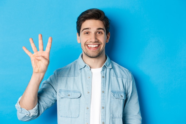 Primer plano de un hombre guapo sonriendo, mostrando los dedos número cuatro, de pie sobre fondo azul.