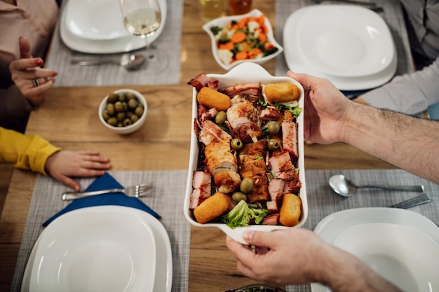 El primer plano de un hombre irreconocible que sirve comida mientras almuerza con su familia en la mesa del comedor