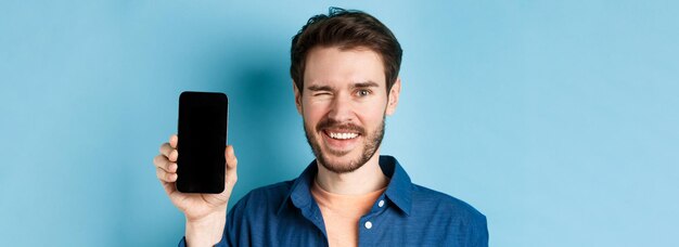 Primer plano de un hombre joven y guapo sonriendo guiñando un ojo y mostrando la pantalla vacía del teléfono móvil de pie en casu