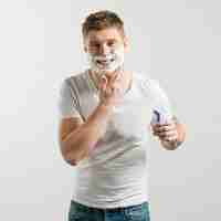 Foto gratuita primer plano de hombre joven sonriente con espuma de afeitar en su cara sosteniendo la maquinilla de afeitar en la mano mirando a la cámara