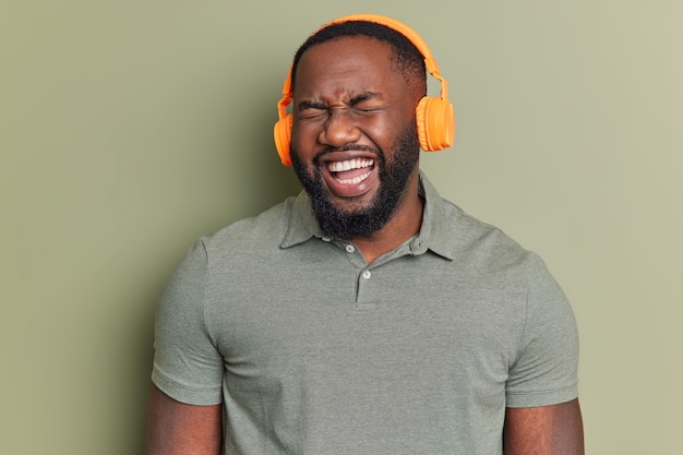 Primer plano de un hombre lleno de alegría con dientes blancos escucha la radio a través de auriculares estéreo oye algo gracioso no puede dejar de reír vestido con una camiseta casual aislada sobre una pared caqui