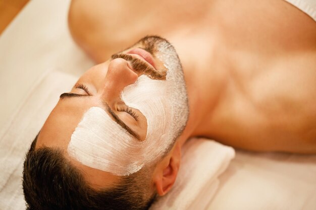 Primer plano de hombre con máscara facial blanca relajante durante el tratamiento en el spa de belleza