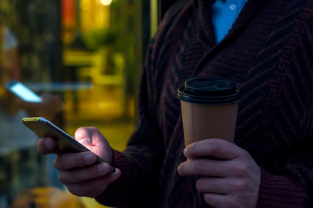 Primer plano de un hombre de negocios usando teléfono móvil y celebración taza de papel. Detalle del primer de una mano de los businessmans que sostiene la taza de papel y que usa un smartphone mientras que recorre en la carretera.