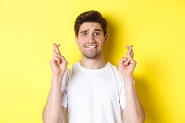 Primer plano de hombre nervioso pidiendo deseos, sosteniendo los dedos cruzados y mordiendo el labio preocupado, de pie sobre fondo amarillo.