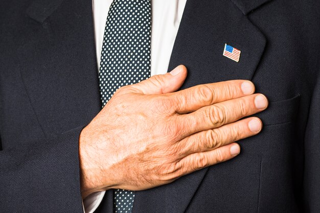 Primer plano de un hombre patriótico con la insignia de Estados Unidos en su abrigo negro tocando la mano en su pecho