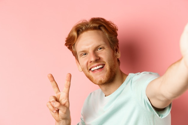 Foto gratuita primer plano de hombre pelirrojo feliz tomando selfie en smartphone y mostrando el signo de la paz, vista desde la cámara del teléfono, fondo rosa.