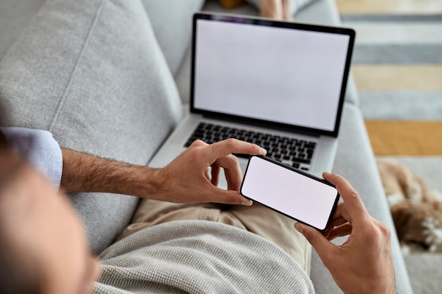 Primer plano de un hombre que usa un teléfono móvil y una computadora portátil mientras se relaja en el sofá El foco está en la pantalla del teléfono inteligente Copiar espacio