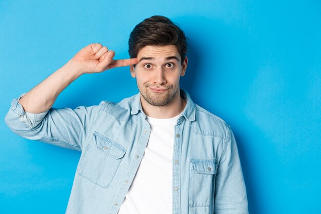 Primer plano del hombre regañando por actuar de forma estúpida o loca, moviendo el dedo en la cabeza y mirando a la cámara, de pie sobre fondo azul