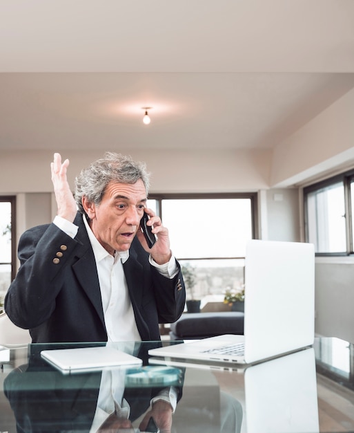 Primer plano de hombre senior mirando portátil hablando por teléfono celular en la oficina