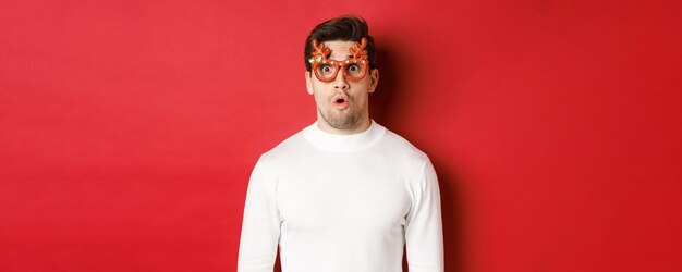 Primer plano de un hombre sorprendido e impresionado con gafas de fiesta de suéter blanco diciendo wow y mirando asombrado