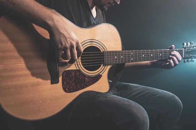 Primer plano de un hombre tocando una guitarra acústica en la oscuridad con iluminación escénica.