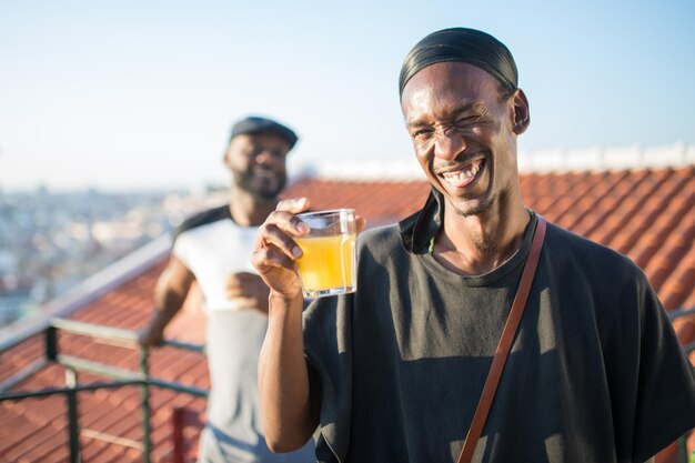 Primer plano de un joven africano con un vaso de jugo en la mano. Un hombre riendo feliz sosteniendo un vaso de jugo de fruta en la cámara y su amigo parado detrás de él. Amistad, concepto de bebidas saludables.