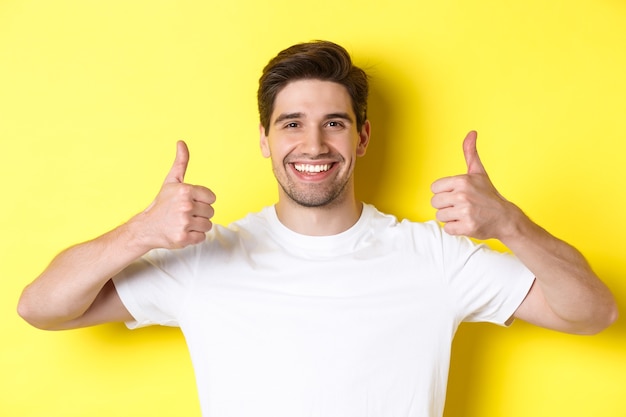 Foto gratuita primer plano de un joven apuesto mostrando los pulgares para arriba, aprobar y estar de acuerdo, sonriendo satisfecho, de pie sobre fondo amarillo.