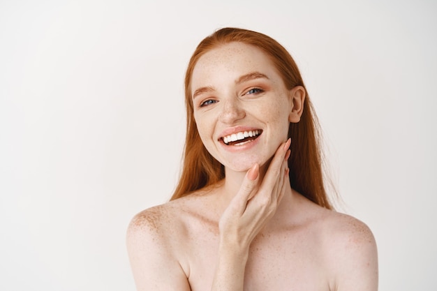 Primer plano de una joven y bella mujer pelirroja sonriendo al frente, tocando la piel limpia perfecta en la cara y luciendo feliz, de pie desnuda sobre una pared blanca