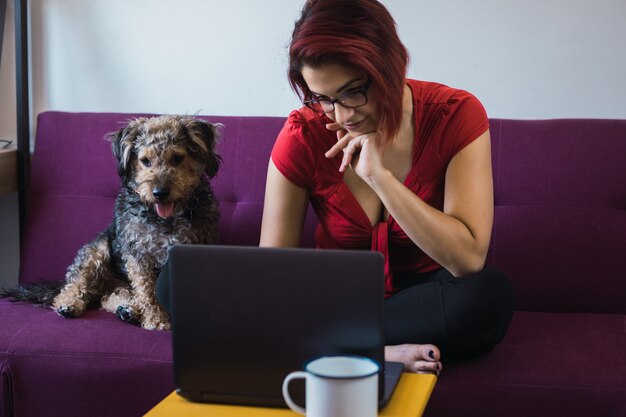 Primer plano de una joven y bella mujer sentada en el sofá con un perro frente a la computadora portátil