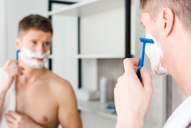 Primer plano de un joven sin camisa afeitado con maquinilla de afeitar azul delante del espejo