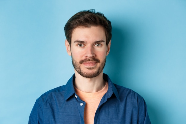 Primer plano de un joven caucásico con barba sonriendo mirando feliz a la cámara, de pie sobre fondo azul.