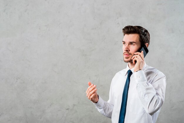 Primer plano de un joven empresario hablando por teléfono inteligente que mira de lejos contra un muro de hormigón gris