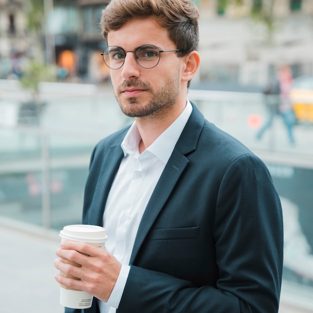 Foto gratuita primer plano de un joven empresario sosteniendo una taza de café disponible en la mano mirando a la cámara