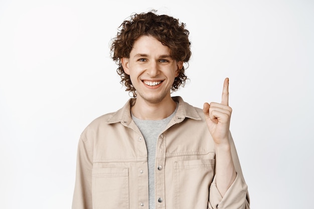 Primer plano de un joven feliz que muestra un anuncio que señala con el dedo hacia arriba de pie sobre fondo blanco