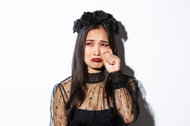 Primer plano de una joven mujer asiática en traje de bruja llorando y mirando miserable, sintiéndose triste, de pie sobre fondo blanco.