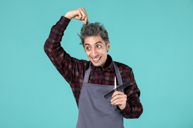 Primer plano de joven peluquero masculino emocional sorprendido vistiendo delantal gris y sosteniendo el peine de tijera tocando el cabello sobre fondo de color azul pastel