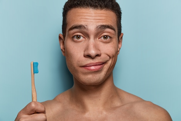 Primer plano de un joven con piel sana, cuerpo fuerte, sostiene el cepillo de dientes, va a someterse a procedimientos higiénicos matutinos, se para contra la pared azul. Concepto de higiene, cuidado dental y belleza.