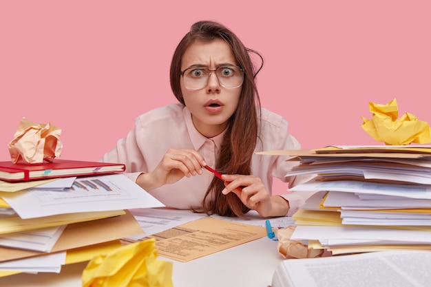 Foto gratuita primer plano de una joven secretaria inteligente disgustada que tiene una expresión de desconcierto, lleva grandes gafas, estudia los documentos y el plazo del contrato