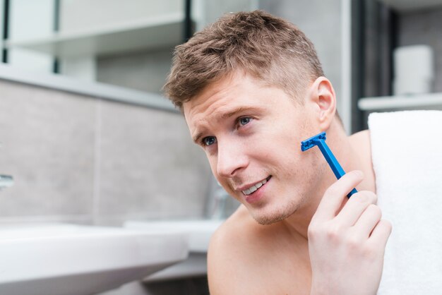 Primer plano de un joven sonriente afeitado con maquinilla de afeitar azul en el baño