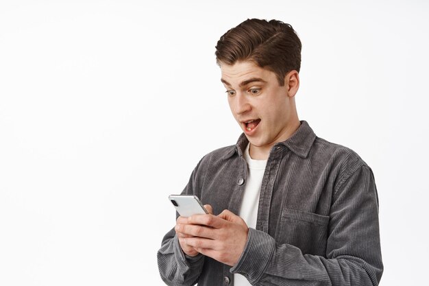 Primer plano de un joven sorprendido mirando el teléfono con asombro, viendo noticias increíbles en el teléfono inteligente, recibiendo la aplicación de notificación, parado sobre fondo blanco