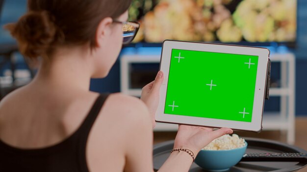 Primer plano de una joven sosteniendo una tableta digital con pantalla verde viendo un seminario web sentado en un sofá frente a la televisión. Chica mirando un dispositivo de pantalla táctil con llave de croma asistiendo a un curso en línea en casa.