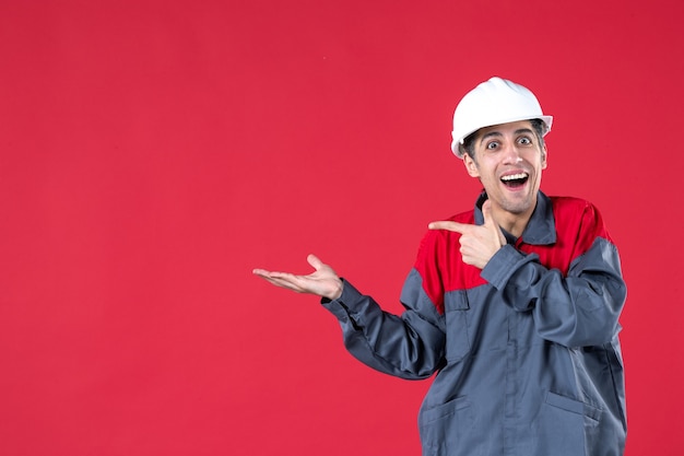 Foto gratuita primer plano de un joven trabajador sorprendido en uniforme con casco apuntando algo en el lado derecho de la pared roja aislada