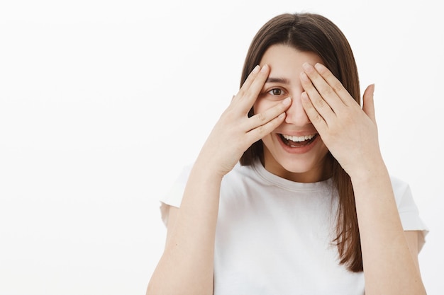 Primer plano de juguetona linda y extrovertida joven morena con camiseta blanca divirtiéndose, riendo y sonriendo mientras juega a escondidas, cerrando los ojos con las palmas de las manos y mirando a través de los dedos alegre