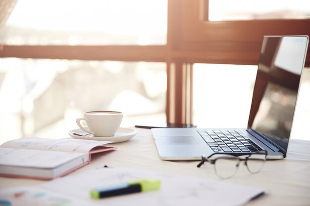 Primer plano lateral de un escritorio de trabajo con la computadora portátil, una taza de café, anteojos y artículos de papelería