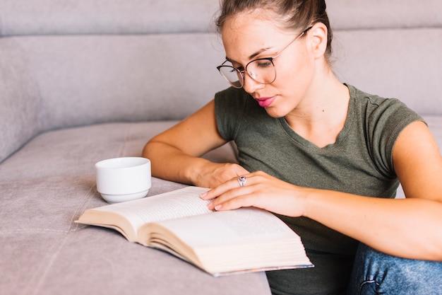 Primer plano de un libro de lectura de mujer joven con una taza de café en el sofá