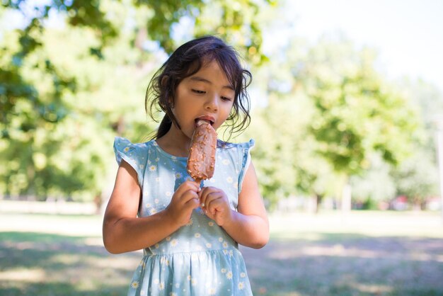 Primer plano de una linda chica asiática disfrutando de un helado en el parque de verano. Niña pequeña con vestido azul parada pensativa con un gran helado en un palo y comiéndola. Concepto de infancia feliz y descanso de verano para niños