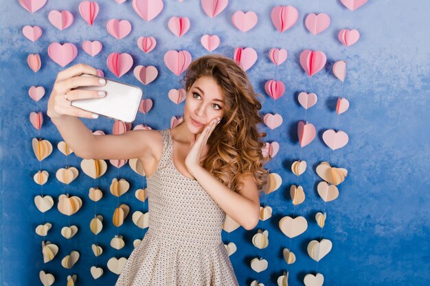 Primer plano de linda chica delgada sexy con pelo rubio rizado de pie en un estudio con fondo azul y escuchando música en auriculares. Ella sonríe, se ve sexy y se hace una selfie.