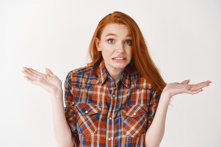 Foto gratuito primer plano de una linda chica pelirroja incómoda encogiéndose de hombros, diciendo ups con cara de culpa y sonrisa confusa, de pie sobre una pared blanca