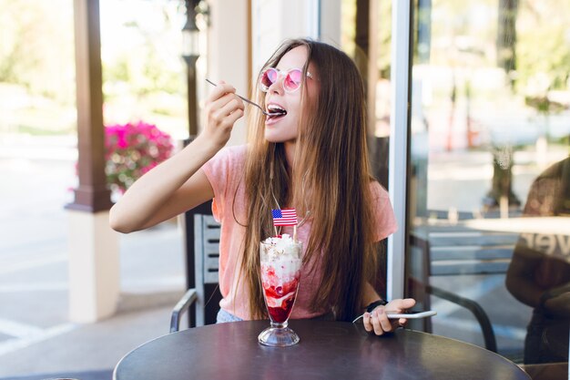 Primer plano de linda chica sentada en un café comiendo helado con cereza en la parte superior con una cuchara. Ella usa top rosa y anteojos rosas. Ella escucha música en su teléfono inteligente. Ella esta disfrutando de su helado