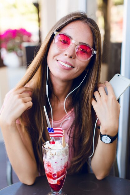 Primer plano de linda chica sentada en un café comiendo helado con cereza en la parte superior. Ella usa top rosa y anteojos rosas. Escucha música en el teléfono inteligente y sonríe. Ella tiene el pelo largo y oscuro