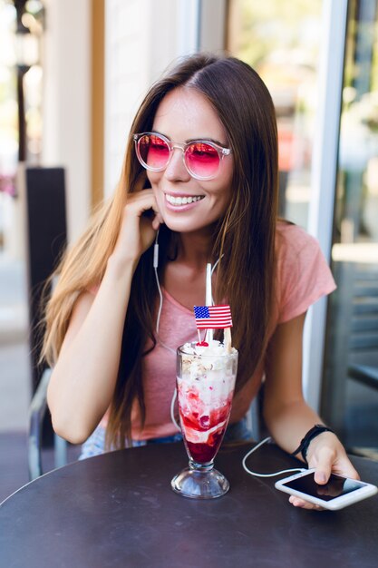 Primer plano de linda chica sentada en un café comiendo helado con cereza en la parte superior. Ella usa top rosa y anteojos rosas. Escucha música en el teléfono inteligente y sonríe. Ella tiene el pelo largo y oscuro
