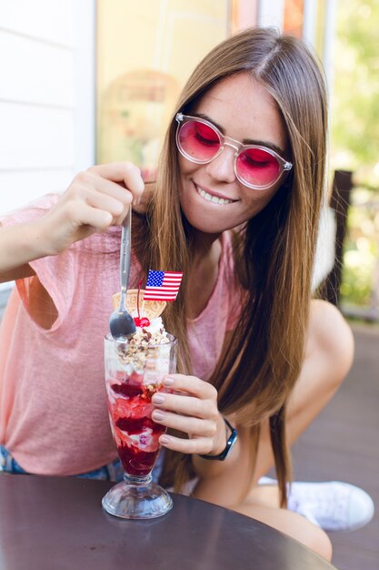 Primer plano de linda chica sentada en una silla comiendo helado con cereza en la parte superior con una cuchara. Viste pantalones cortos de mezclilla, top rosa y sonríe. Ella tiene anteojos rosas