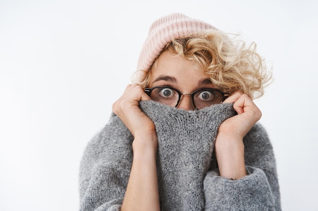 Primer plano de una linda chica sorprendida y sin palabras en un gorro de invierno y gafas tirando del cuello del suéter en la cara del miedo que estalla los ojos