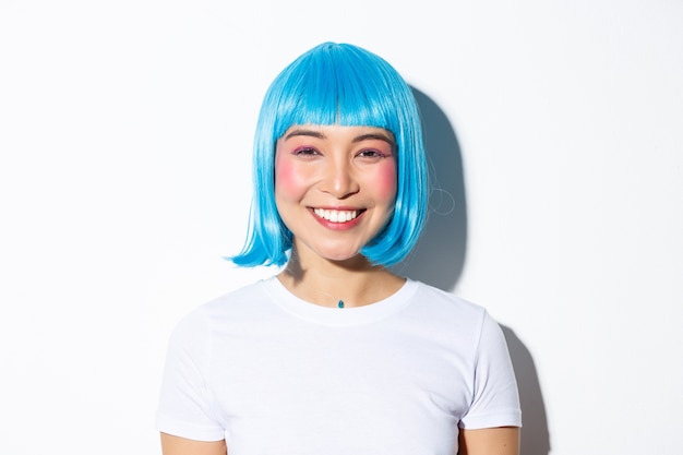 Primer plano de linda mujer asiática celebrando halloween con peluca azul, sonriendo feliz a la cámara, de pie.
