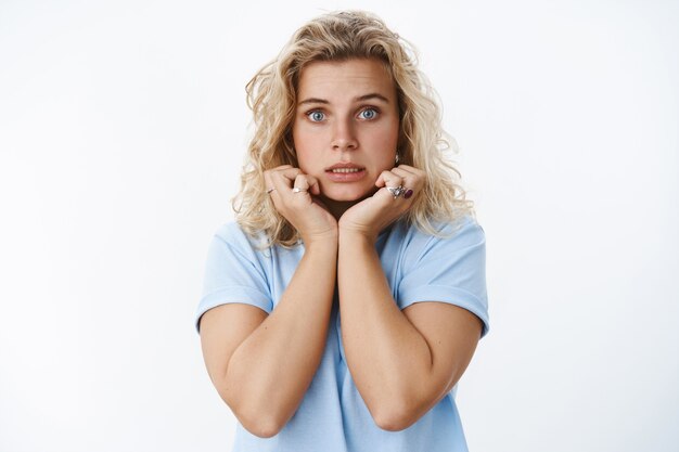 Primer plano de una linda mujer insegura y tonta asustada con cabello rubio y ojos azules aprieta los dientes temblando de miedo tomados de la mano en la línea de la mandíbula, preocupada por estar preocupada y confundida, expresando nerviosismo